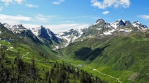A picturesque mountainous landscape featuring snow-capped peaks under a clear blue sky. Verdant valleys with lush greenery stretch between the mountains, winding roads, and scattered pine trees add to the natural beauty. A few small structures are visible in the valley.