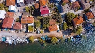 An aerial view displays a coastal neighborhood with houses featuring red-tiled roofs. The shoreline is lined with small boats and a rocky edge, while trees and greenery are interspersed among the homes. Streets and pathways connect the buildings, and vehicles can be seen parked along the streets.
