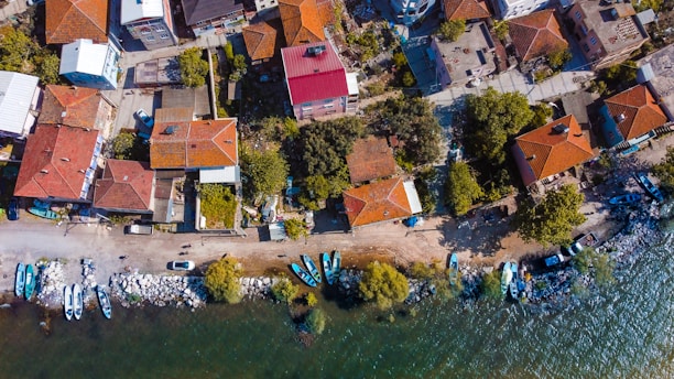 An aerial view displays a coastal neighborhood with houses featuring red-tiled roofs. The shoreline is lined with small boats and a rocky edge, while trees and greenery are interspersed among the homes. Streets and pathways connect the buildings, and vehicles can be seen parked along the streets.