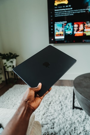 A hand is holding a black Apple laptop in a modern living room. In the background, a large television displays a streaming service interface with various movie thumbnails. The room features a gray shaggy carpet, a dark round coffee table, and a plant in the corner.