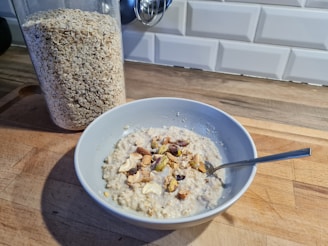 A bowl of oatmeal topped with various nuts is placed on a wooden kitchen counter. Next to it is a transparent container filled with rolled oats. The background features a white tile backsplash.