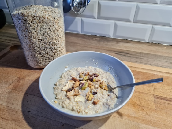 A bowl of oatmeal topped with various nuts is placed on a wooden kitchen counter. Next to it is a transparent container filled with rolled oats. The background features a white tile backsplash.