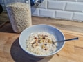 A bowl of oatmeal topped with various nuts is placed on a wooden kitchen counter. Next to it is a transparent container filled with rolled oats. The background features a white tile backsplash.