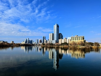 A skyline featuring modern skyscrapers and buildings reflecting in a calm body of water, under a bright blue sky filled with scattered clouds.