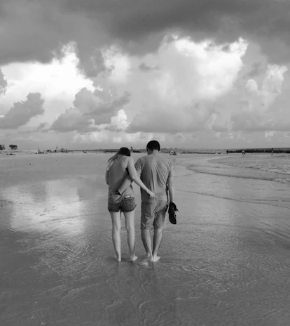 A candid shot of a couple holding hands, walking barefoot along a sun-drenched shoreline.
