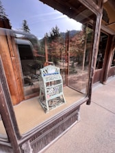 A vintage-style decorative object with intricate metalwork sits in a glass display case. The object appears to be an ornate lantern or metal piece with cut-out patterns. It is placed on a tatami mat against a wooden backdrop with exterior reflections visible in the glass, showcasing trees and people outside.