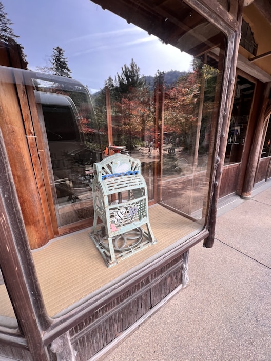 A vintage-style decorative object with intricate metalwork sits in a glass display case. The object appears to be an ornate lantern or metal piece with cut-out patterns. It is placed on a tatami mat against a wooden backdrop with exterior reflections visible in the glass, showcasing trees and people outside.