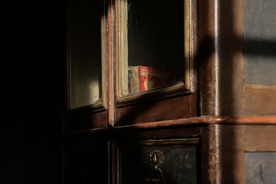 A wooden cabinet with glass panels displaying books partially obscured in dim lighting. Sunlight casts shadows across the surface, highlighting the textures of the wood.
