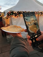 A hand holding a plate with a crepe in front of a festive tent adorned with holiday lights and decorations. Another hand holds a smartphone, capturing a photo of the crepe with a ferris wheel visible on the phone screen.