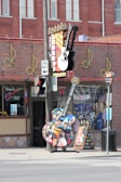 A vibrant city street corner known as Legends Corner, featuring a large guitar-shaped sign adorned with images of famous musicians. The brick building is decorated with musical notes and displays signs for live music venues. A large, colorful guitar cutout and a billboard are positioned outside.