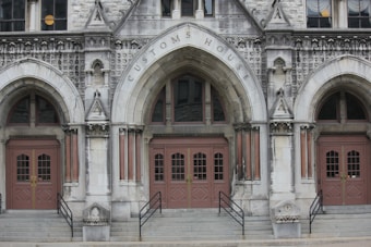 An old, ornate stone building with three arched entrances labeled 'Customs House' above the main arch. The doors are wooden with glass panels, surrounded by detailed carvings and columns. Decorative stonework adorns the facade.