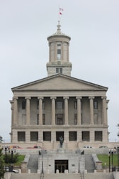 A neoclassical government building with a prominent tower and flag flying at the top. It features multiple columns on its facade and a central statue at the front, with steps leading up to the entrance. Several vehicles are parked in front of the building.