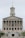 A neoclassical government building with a prominent tower and flag flying at the top. It features multiple columns on its facade and a central statue at the front, with steps leading up to the entrance. Several vehicles are parked in front of the building.