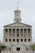 A neoclassical government building with a prominent tower and flag flying at the top. It features multiple columns on its facade and a central statue at the front, with steps leading up to the entrance. Several vehicles are parked in front of the building.