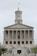 A neoclassical government building with a prominent tower and flag flying at the top. It features multiple columns on its facade and a central statue at the front, with steps leading up to the entrance. Several vehicles are parked in front of the building.