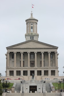 A neoclassical government building with a prominent tower and flag flying at the top. It features multiple columns on its facade and a central statue at the front, with steps leading up to the entrance. Several vehicles are parked in front of the building.