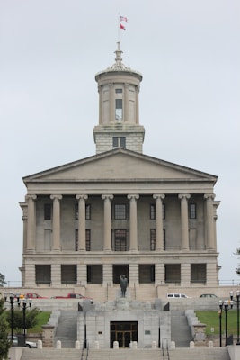 A neoclassical government building with a prominent tower and flag flying at the top. It features multiple columns on its facade and a central statue at the front, with steps leading up to the entrance. Several vehicles are parked in front of the building.