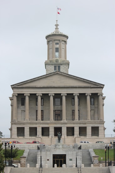 A neoclassical government building with a prominent tower and flag flying at the top. It features multiple columns on its facade and a central statue at the front, with steps leading up to the entrance. Several vehicles are parked in front of the building.