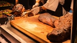 A person wearing a white shirt and gloves is slicing a large piece of roasted meat on a wooden cutting board. The meat has a crispy, seasoned crust and is surrounded by various dishes and garnishes, including colorful salads and berries.
