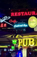 Colorful neon signs are prominently displayed, advertising a restaurant, cafe, pub, cocktail bar, and pizzeria. Various logo designs and typefaces are used, featuring brands like Pilsner Urquell and Jim Beam. The signs are illuminated against a dark background, creating a lively and vibrant atmosphere.