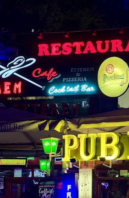 Colorful neon signs are prominently displayed, advertising a restaurant, cafe, pub, cocktail bar, and pizzeria. Various logo designs and typefaces are used, featuring brands like Pilsner Urquell and Jim Beam. The signs are illuminated against a dark background, creating a lively and vibrant atmosphere.