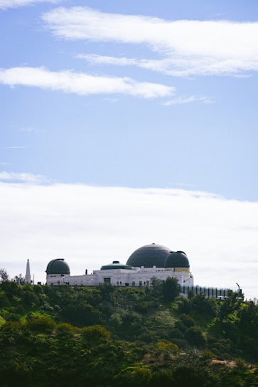 An observatory with a large white structure featuring multiple domes sits atop a green hill. The sky is clear with some scattered clouds, creating a contrast with the lush vegetation surrounding the building.