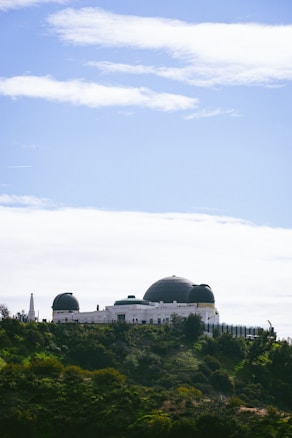 An observatory with a large white structure featuring multiple domes sits atop a green hill. The sky is clear with some scattered clouds, creating a contrast with the lush vegetation surrounding the building.