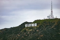 A large, iconic white sign displaying the word 'Hollywood' is situated on a lush, green hillside. An extensive array of telecommunications antennas and towers is located to the right on the hilltop, contrasting with the cloudy, overcast sky above.