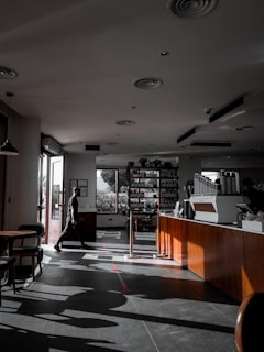 A modern coffee shop with dark flooring and wooden furniture. Sunlight streams through large windows, casting shadows and highlighting an angled design across the interior. A person walks near the entrance, which is open, allowing natural light to flood the space. Shelves and a counter with a coffee machine can be seen, adding to the cozy ambiance.