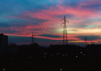 Silhouettes of power lines and towers stand against a vibrant sunset sky featuring shades of pink, purple, and blue. The foreground is dark, creating a striking contrast with the colorful horizon.