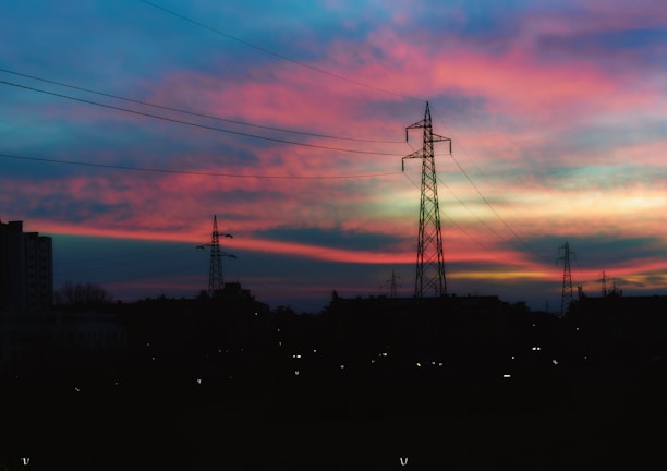 Silhouettes of power lines and towers stand against a vibrant sunset sky featuring shades of pink, purple, and blue. The foreground is dark, creating a striking contrast with the colorful horizon.