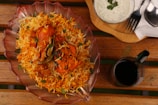 A richly colored dish of chicken biryani is presented in a decorative glass bowl on a wooden table. The biryani is garnished with fresh coriander leaves and green chilies, exuding vibrant orange and yellow hues. Accompanied by a bowl of raita topped with herbs and a glass of dark-colored beverage. Utensils are placed neatly on a wooden tray beside the dish.