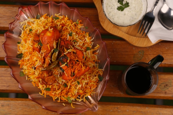 A richly colored dish of chicken biryani is presented in a decorative glass bowl on a wooden table. The biryani is garnished with fresh coriander leaves and green chilies, exuding vibrant orange and yellow hues. Accompanied by a bowl of raita topped with herbs and a glass of dark-colored beverage. Utensils are placed neatly on a wooden tray beside the dish.