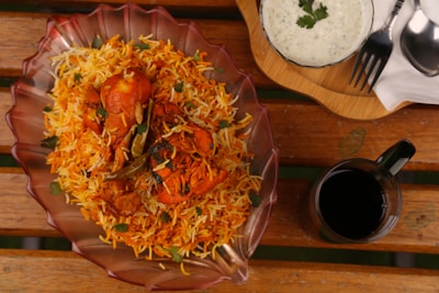A richly colored dish of chicken biryani is presented in a decorative glass bowl on a wooden table. The biryani is garnished with fresh coriander leaves and green chilies, exuding vibrant orange and yellow hues. Accompanied by a bowl of raita topped with herbs and a glass of dark-colored beverage. Utensils are placed neatly on a wooden tray beside the dish.