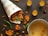 A newspaper cone filled with crispy brown fritters is placed on a dark surface, surrounded by green chili peppers. Nearby, a metal pot with remnants of sauce sits next to three small glass containers filled with a yellow liquid, arranged neatly.