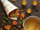 A newspaper cone filled with crispy brown fritters is placed on a dark surface, surrounded by green chili peppers. Nearby, a metal pot with remnants of sauce sits next to three small glass containers filled with a yellow liquid, arranged neatly.