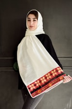 A person wearing a white headscarf with an intricate red and orange floral border pattern. They are dressed in a dark top and standing against a plain dark background. The headscarf drapes down across their front.