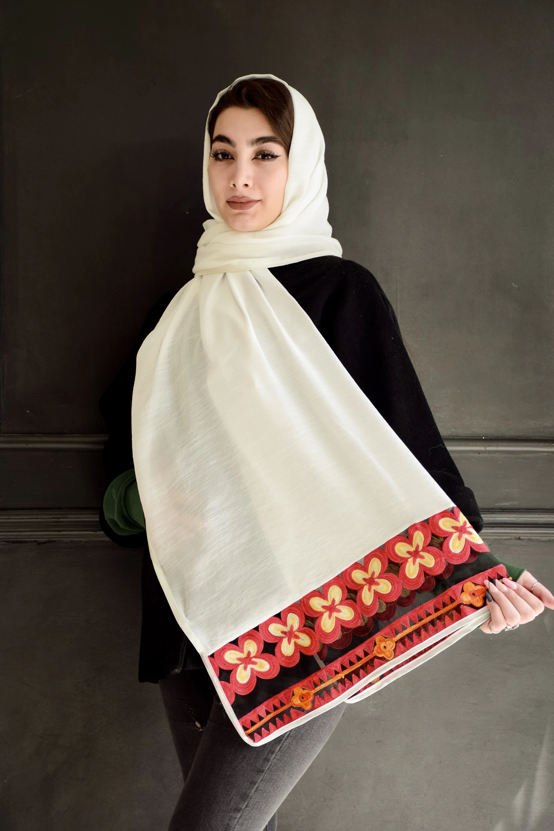 A person wearing a white headscarf with an intricate red and orange floral border pattern. They are dressed in a dark top and standing against a plain dark background. The headscarf drapes down across their front.