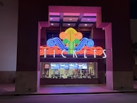 A colorful neon sign with swirling designs in red, blue, green, and yellow is prominently displayed above a ticket counter. The word 'TICKETS' is illuminated in white letters below the swirling design. Behind the glass window, tickets and posters for movies are visible, suggesting a theater or cinema setting.