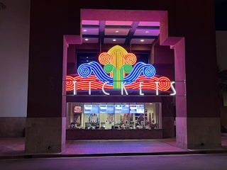 A colorful neon sign with swirling designs in red, blue, green, and yellow is prominently displayed above a ticket counter. The word 'TICKETS' is illuminated in white letters below the swirling design. Behind the glass window, tickets and posters for movies are visible, suggesting a theater or cinema setting.