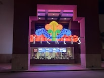 A colorful neon sign with swirling designs in red, blue, green, and yellow is prominently displayed above a ticket counter. The word 'TICKETS' is illuminated in white letters below the swirling design. Behind the glass window, tickets and posters for movies are visible, suggesting a theater or cinema setting.
