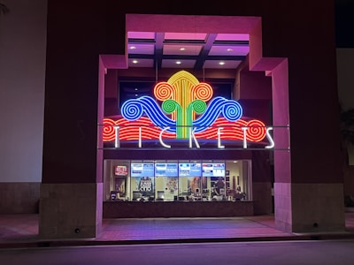 A colorful neon sign with swirling designs in red, blue, green, and yellow is prominently displayed above a ticket counter. The word 'TICKETS' is illuminated in white letters below the swirling design. Behind the glass window, tickets and posters for movies are visible, suggesting a theater or cinema setting.