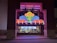 A colorful neon sign with swirling designs in red, blue, green, and yellow is prominently displayed above a ticket counter. The word 'TICKETS' is illuminated in white letters below the swirling design. Behind the glass window, tickets and posters for movies are visible, suggesting a theater or cinema setting.