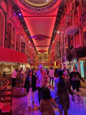 A vibrant, indoor area with a walkway lined by shops or displays. The ceiling is illuminated with red and pink lighting, creating a lively atmosphere. People dressed in casual attire, including tropical shirts, are walking and talking. The setting appears to be festive or part of an entertainment complex.