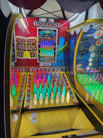 An arcade ring toss game featuring rows of brightly colored bottles under a canopy with lights. The setup includes a variety of colored bottles arranged in a neat grid, with a scoreboard displaying points and bonuses hanging on the back panel. The game is enclosed in a frame with vibrant graphics and instructions.