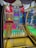 An arcade ring toss game featuring rows of brightly colored bottles under a canopy with lights. The setup includes a variety of colored bottles arranged in a neat grid, with a scoreboard displaying points and bonuses hanging on the back panel. The game is enclosed in a frame with vibrant graphics and instructions.