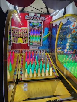 An arcade ring toss game featuring rows of brightly colored bottles under a canopy with lights. The setup includes a variety of colored bottles arranged in a neat grid, with a scoreboard displaying points and bonuses hanging on the back panel. The game is enclosed in a frame with vibrant graphics and instructions.