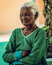 An elderly person with white hair is wearing a green blouse with gold trim, seated outdoors in front of a blurred background with a tree on the side. Their expression appears contemplative.