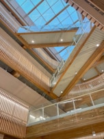 A modern architectural interior featuring a series of wooden beams and glass railings. The skylight ceiling allows natural light to flood the space, highlighting the clean lines and geometric design elements. The wood tones add warmth and contrast with the sleekness of the glass and metal.