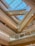 A modern architectural interior featuring a series of wooden beams and glass railings. The skylight ceiling allows natural light to flood the space, highlighting the clean lines and geometric design elements. The wood tones add warmth and contrast with the sleekness of the glass and metal.